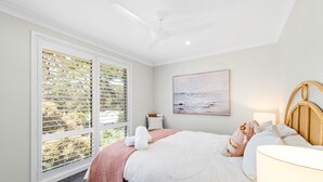 A bedroom with a bed, a ceiling fan, a window with blinds, a painting on the wall, and a bedside table with a lamp. - Bonito near Bagnalls Beach Corlette (Corlette)