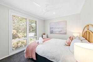 Een slaapkamer met een bed, een plafondventilator, een raam met zonwering, een schilderij aan de muur en een nachtkastje met een lamp.