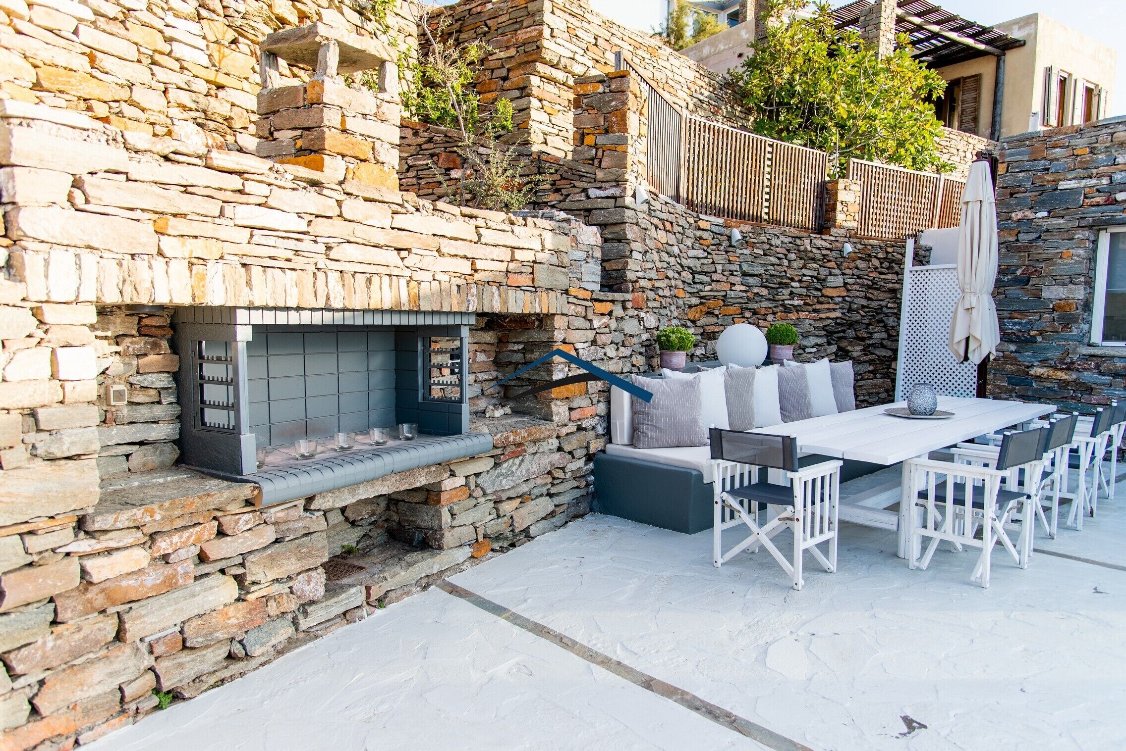 A stone-walled outdoor area with a built-in grill, a white table with chairs, and a pergola with a white umbrella.