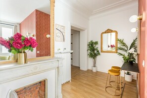 A room with a white fireplace, a mirror, a door with a red panel, a plant, a table with two yellow stools, and a mirror with a gold frame. - Merveil - Family Suite - Opera - Cardinal Mercier (Paris)