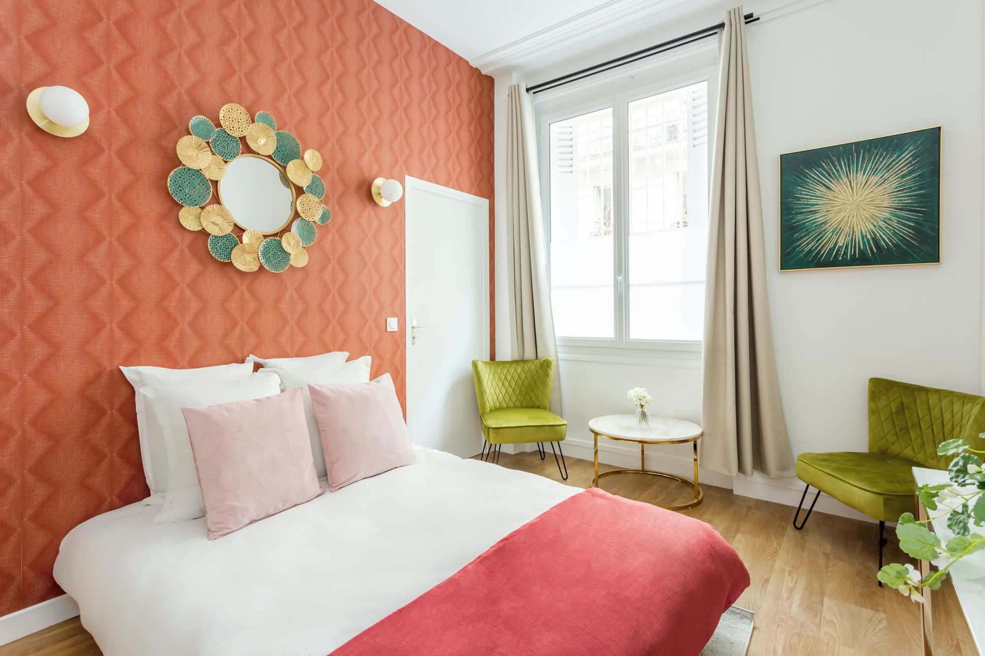 A bedroom with a bed, two chairs, a small table, a mirror, and a window with curtains.