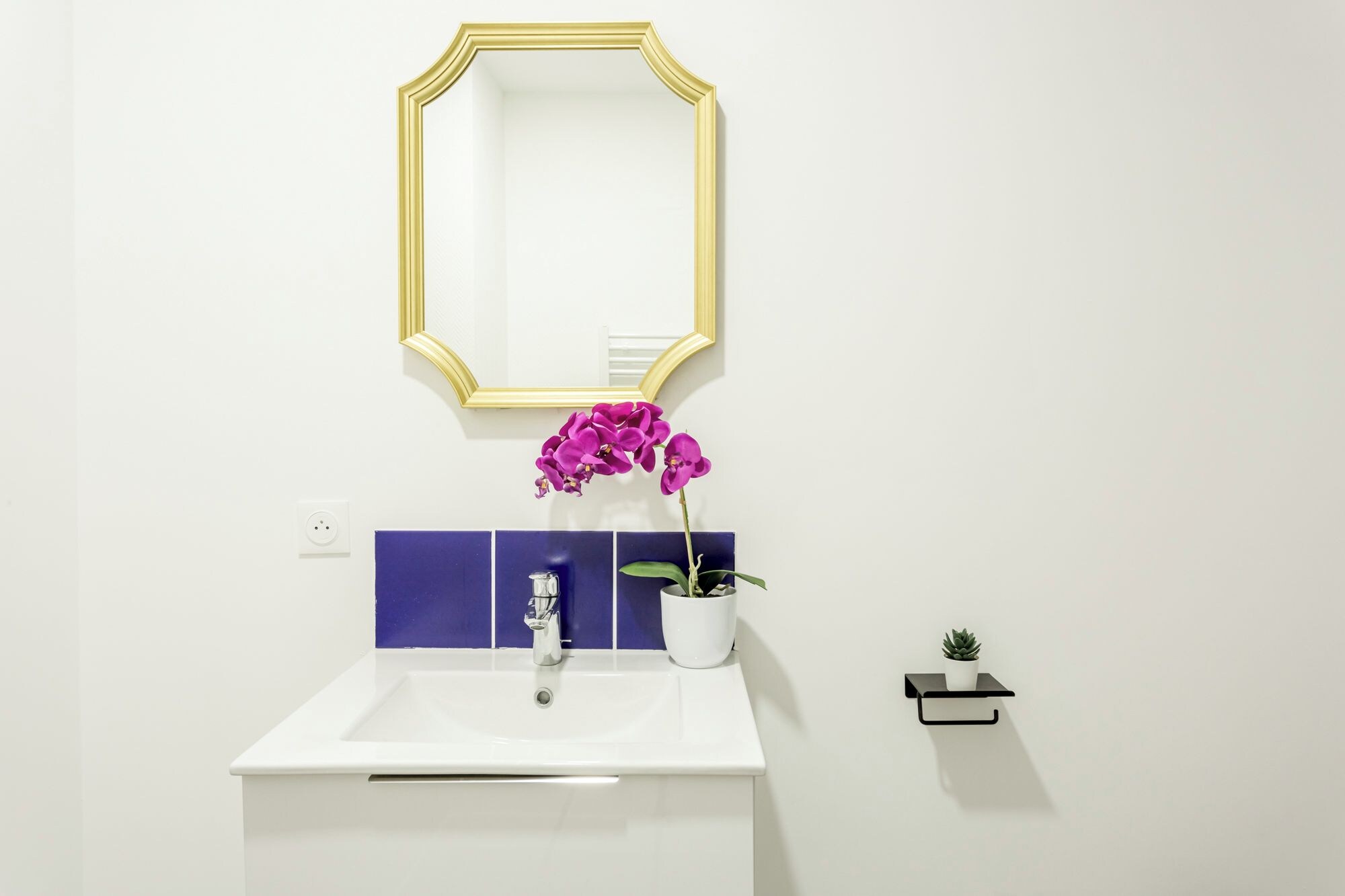 Ein Badezimmer mit einem weißen Waschbecken, einem Spiegel mit goldener Rahmung, lila Blumen und einer kleinen Topfpflanze.
