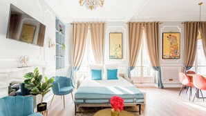 A well-lit room with a bed, a blue armchair, a small table with a vase, and a red chair. - Merveil - Luxury Suite - Opera - Septembre I (Paris)