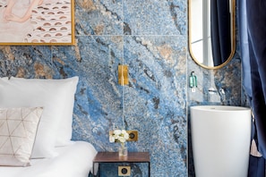 A bedroom with a bed, a nightstand with a vase of flowers, a wall-mounted light switch, and a bathroom with a sink and mirror. - Merveil - Luxury Suite - Opera - Septembre I (Paris)