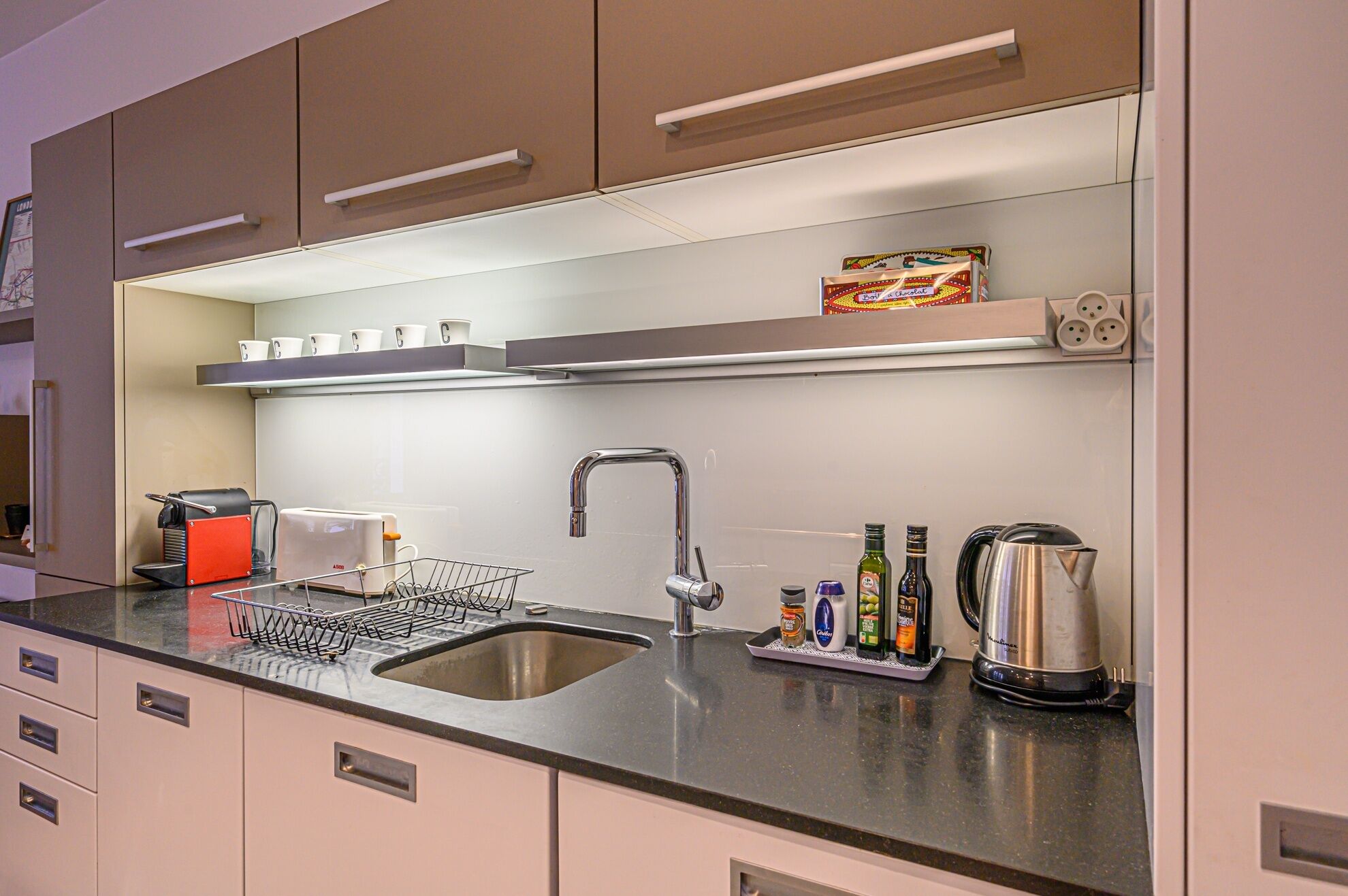 A modern kitchen with a stainless steel sink, a coffee machine, a kettle, and a shelf with cups and a box of chocolate.