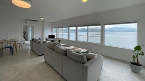 A spacious living area with a large window overlooking a body of water, a sofa, a dining table, and a television.