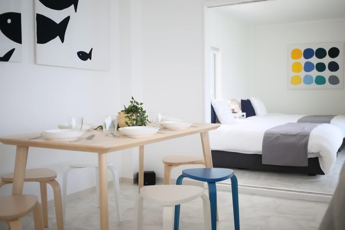 Une chambre d’hôtel moderne avec une table à manger en bois, des chaises blanches, un lit aux couleurs gris et blanc, et un mur orné d’œuvres d’art abstraites.