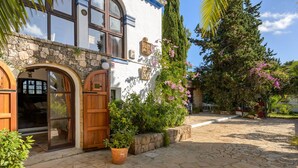 A courtyard with stone flooring, a wooden door, and a white building with stone walls and arched windows. - Villa Can Sarrio - San Carlos (Santa Eulària des Riu)