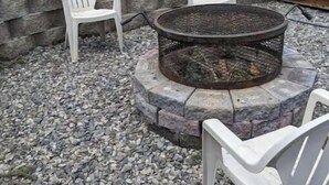 Un hogar de piedra con dos sillas de plástico blanco a su alrededor, sobre una superficie de grava y con un muro de piedra al fondo.