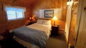 Un dormitorio acogedor tipo cabaña de madera con una cama, dos lámparas de noche, una ventana con persianas, un cuadro en la pared y una puerta.