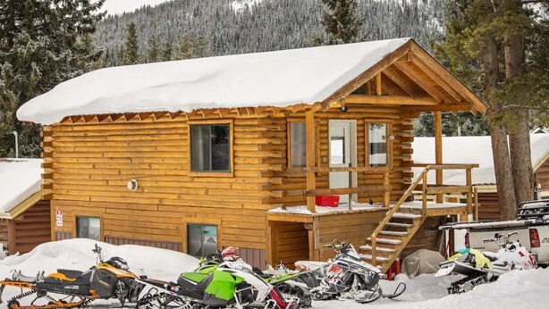 A wooden cabin with a snow-covered roof, a staircase leading to a deck, and several snowmobiles parked in front. - Eagle Nest Cabin Yellowstone Getaway in Cooke City (Cooke city)