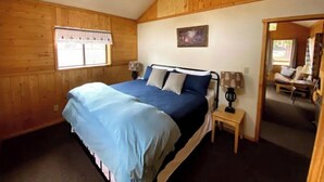 A bedroom with a bed, a nightstand, a lamp, a window with a curtain, and a painting on the wall. - Eagle Nest Cabin Yellowstone Getaway in Cooke City (Cooke city)