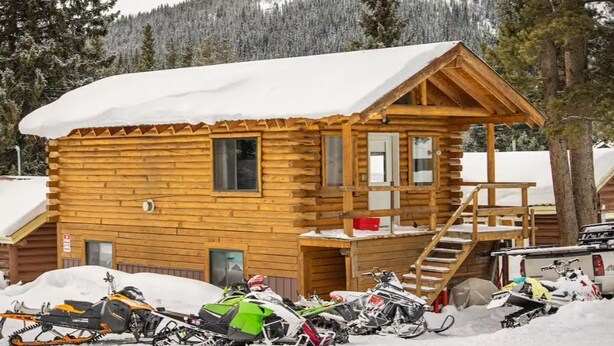 A wooden cabin with a snow-covered roof, a front porch, and a staircase leading to the entrance. Several snowmobiles are parked in front.