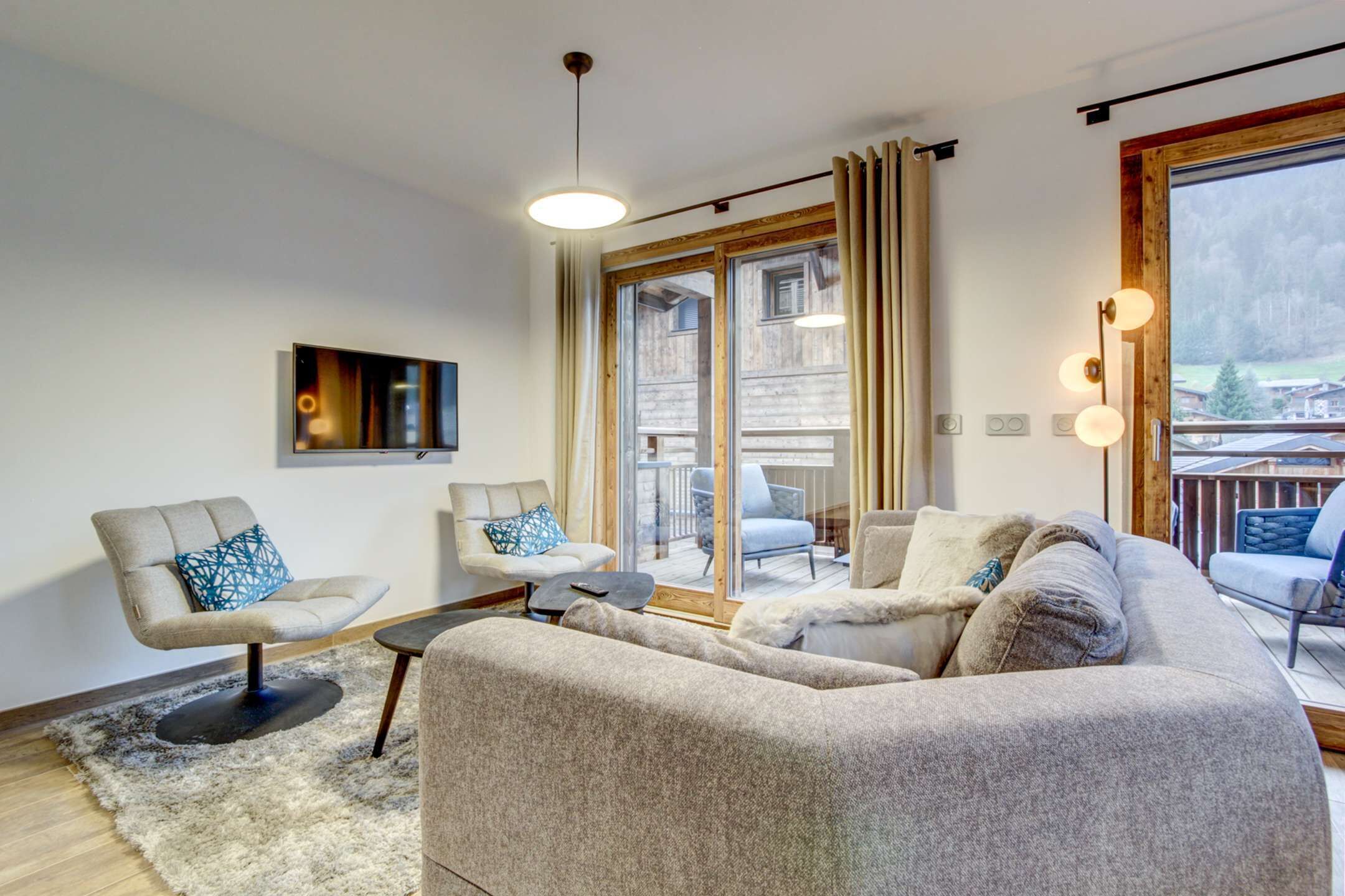 Una sala de estar moderna con sofá, sillones y una mesa de centro. Hay un ventanal con cortinas, una lámpara de techo y vistas a una terraza y edificios al exterior.