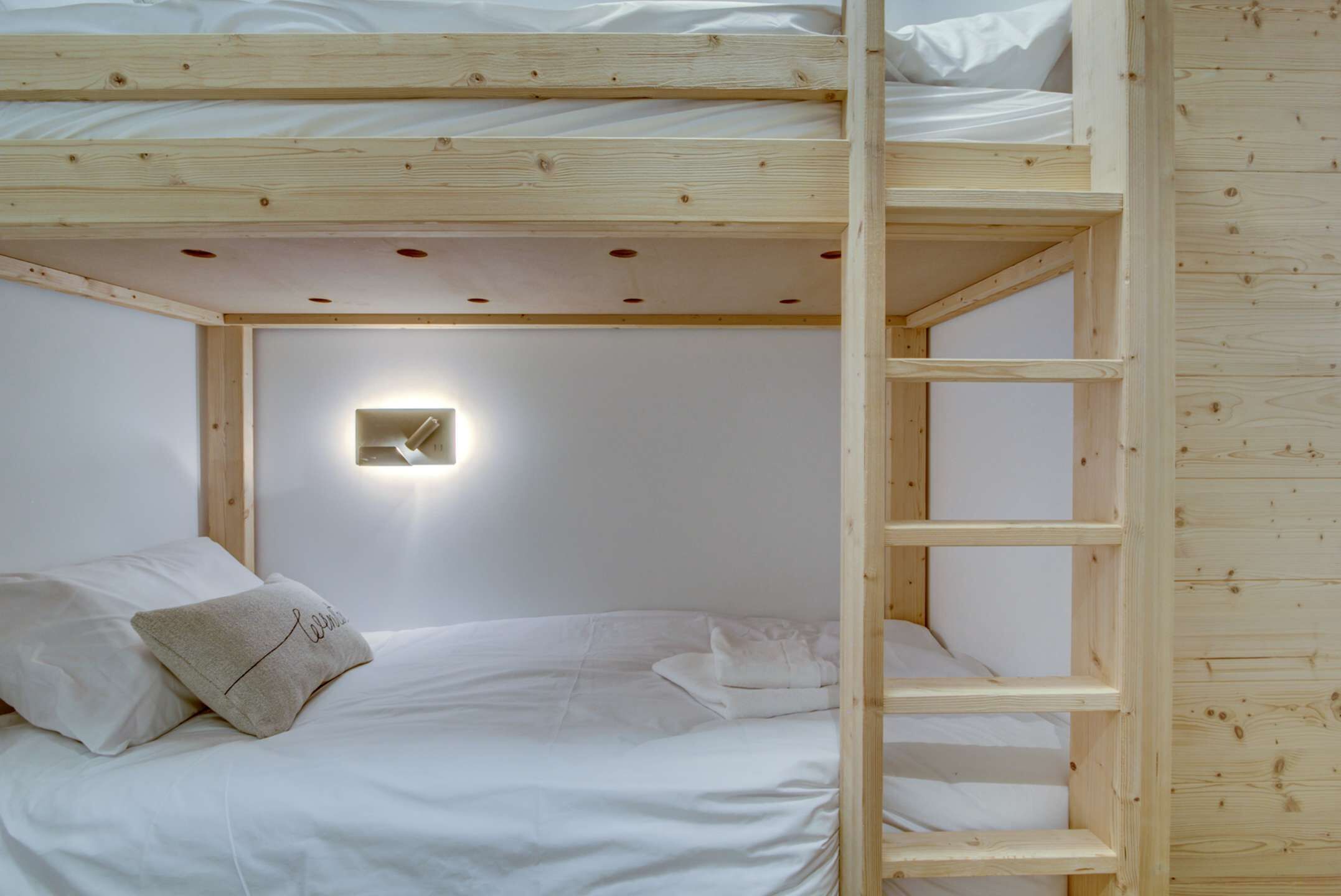 Een slaapkamer met een stapelbed, een ladder, wit beddengoed en een wandlamp.