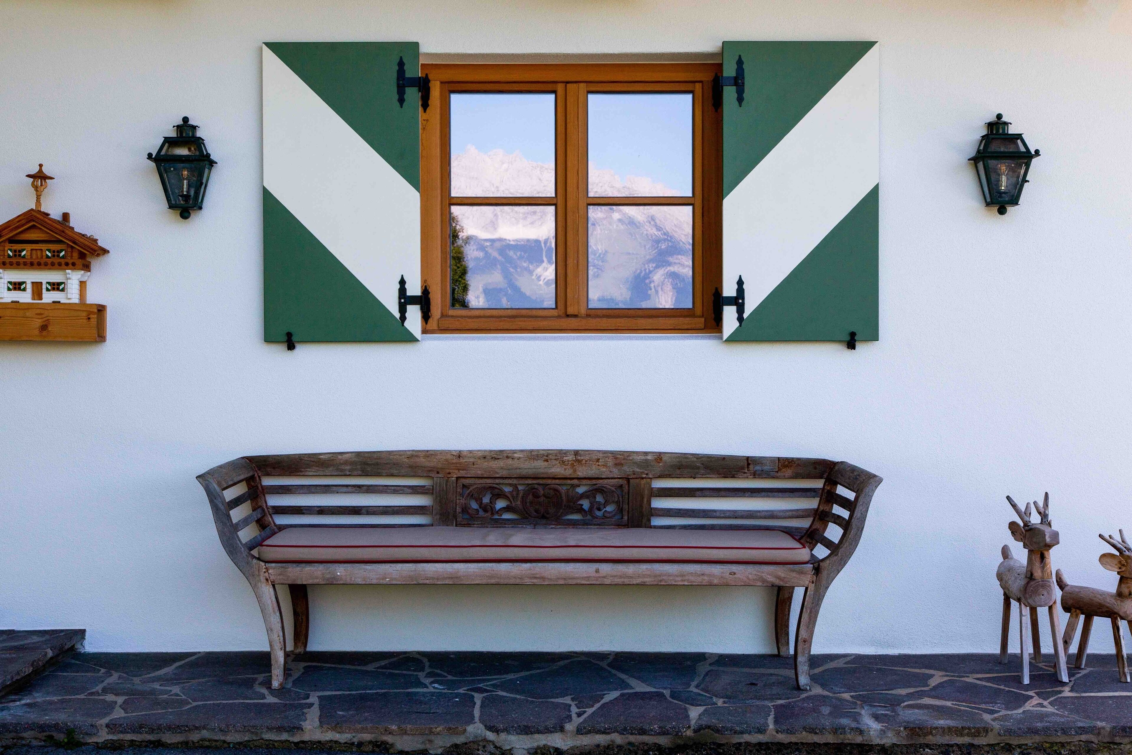 A wooden bench with a view of mountains through a window with green shutters.