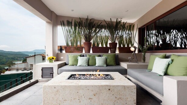 Un área de estar al aire libre moderna con una chimenea, un sofá con cojines verdes y plantas altas en macetas.