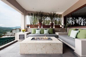A modern outdoor seating area with a fire pit, a sofa with green cushions, and tall potted plants.