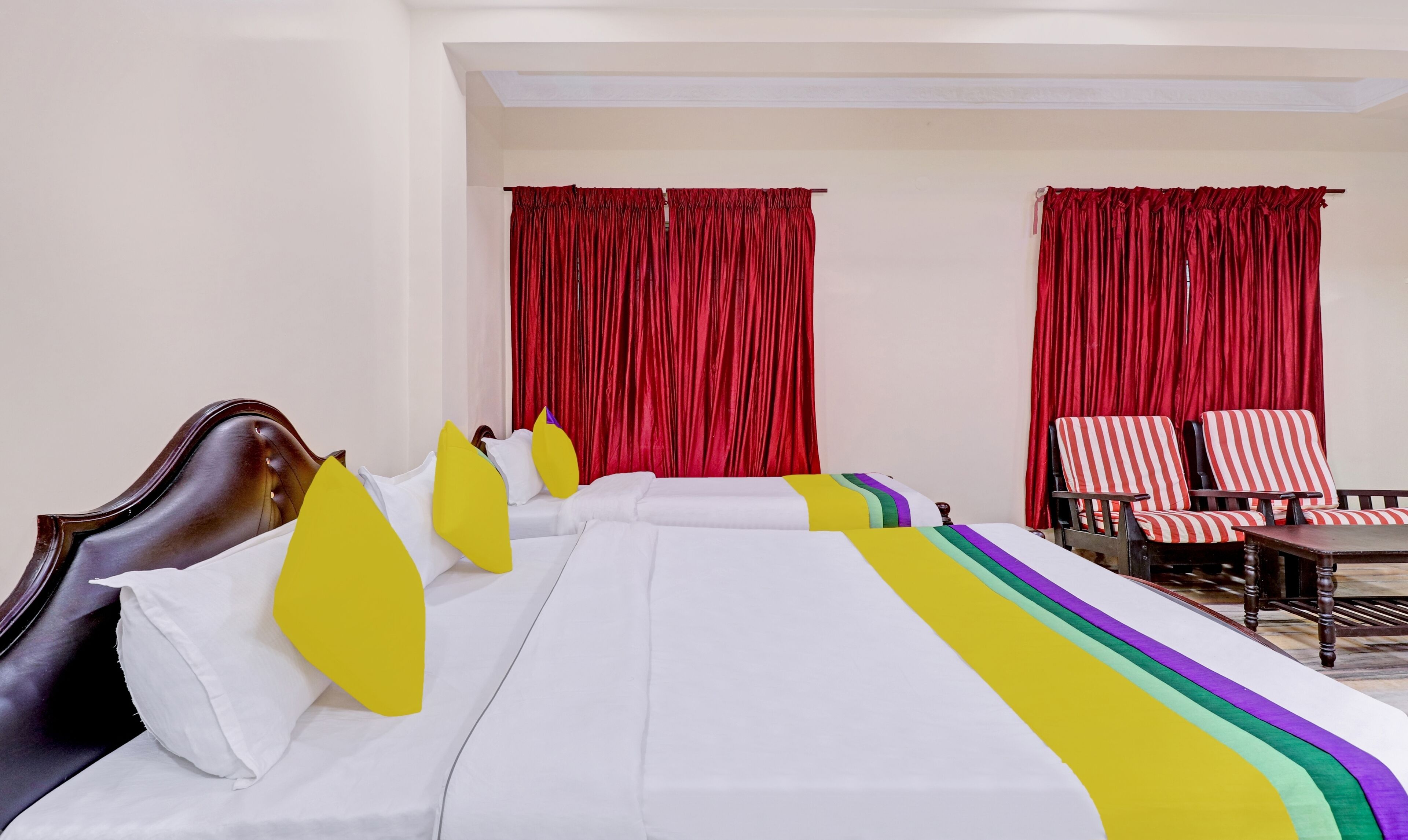 A hotel room with a large bed, two bedside tables, a wooden headboard, red curtains, striped chairs, and a small table.