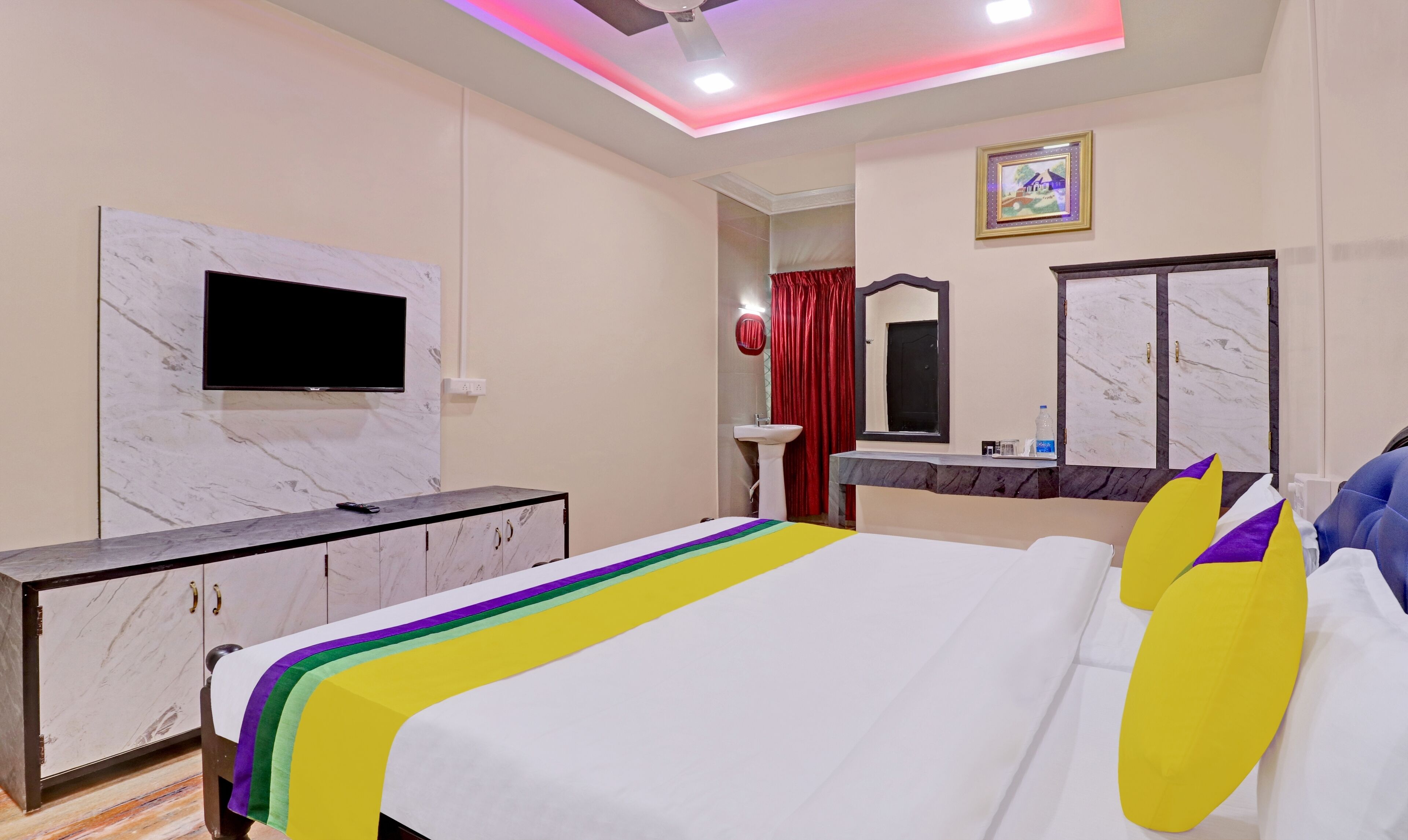 A hotel room with a bed, a TV mounted on the wall, a vanity with a mirror, and a ceiling fan.
