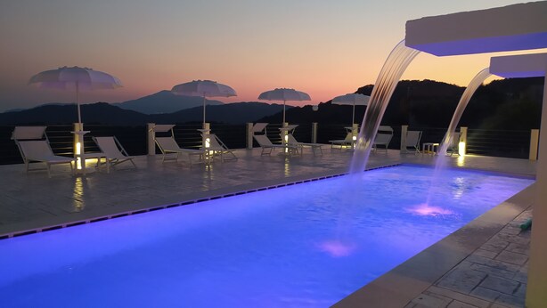 Zona de piscina con sillones reclinables y sombrillas, una fuente de agua y vistas a montañas al fondo.