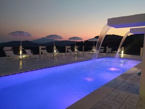 A poolside area with lounge chairs and umbrellas, a water feature, and a view of mountains in the background. - Tenuta Vallina (Tortorella)