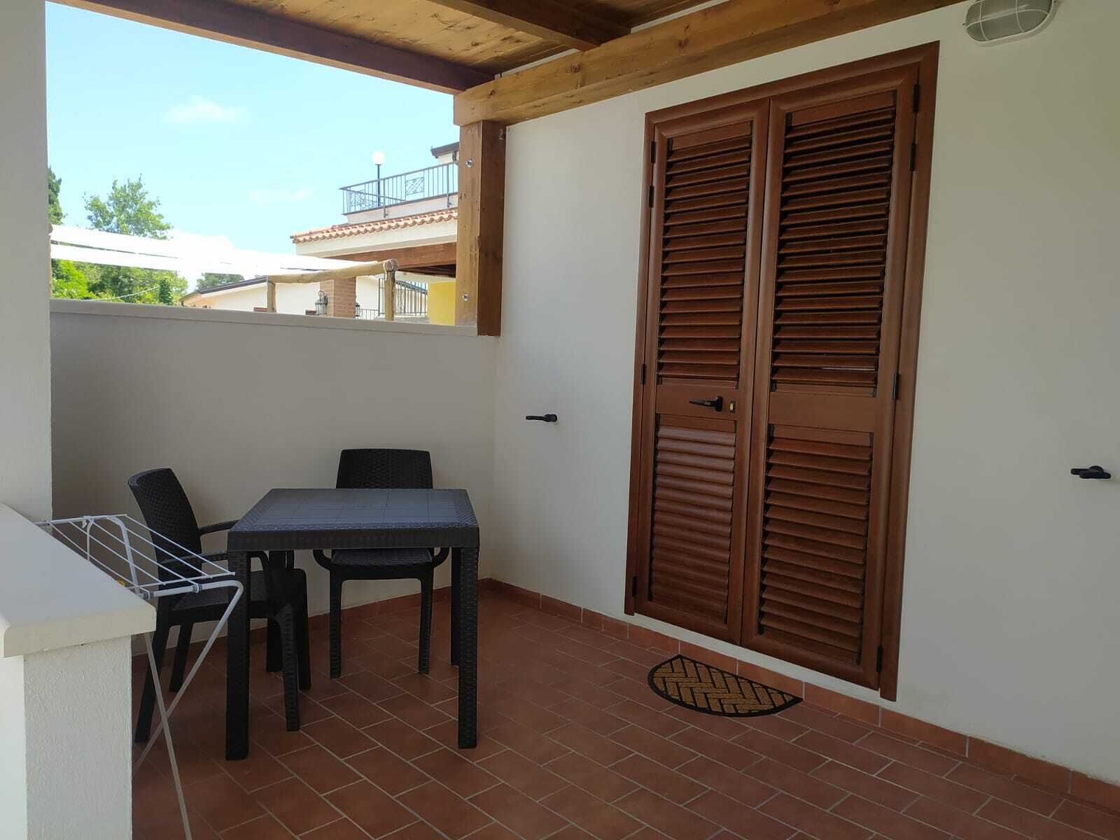 Un balcone con tavolo e sedie, una porta in legno e vista su un edificio sullo sfondo.