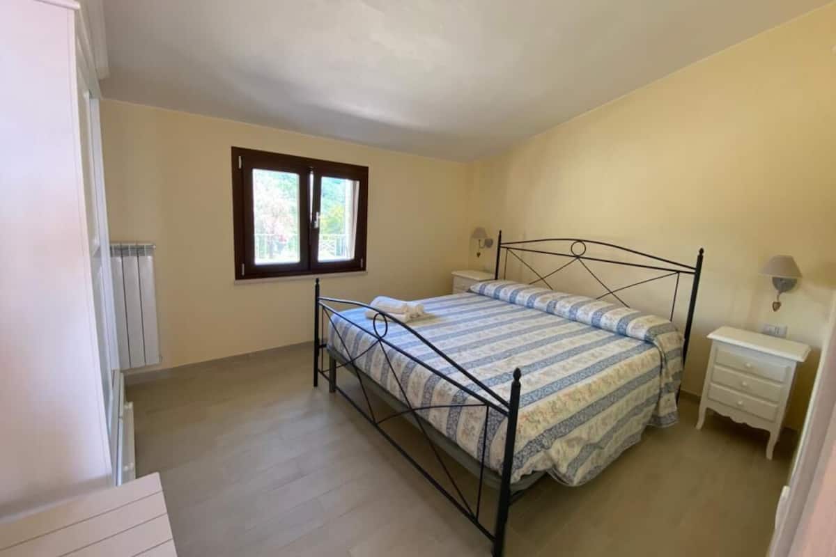 A bedroom with a bed, a nightstand, a window, and a radiator.