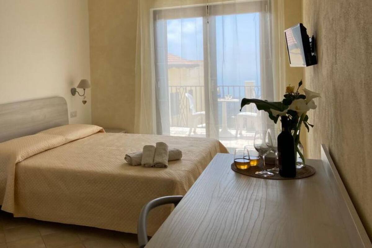 A hotel room with a bed, bedside table, lamp, and a view of the outside balcony.