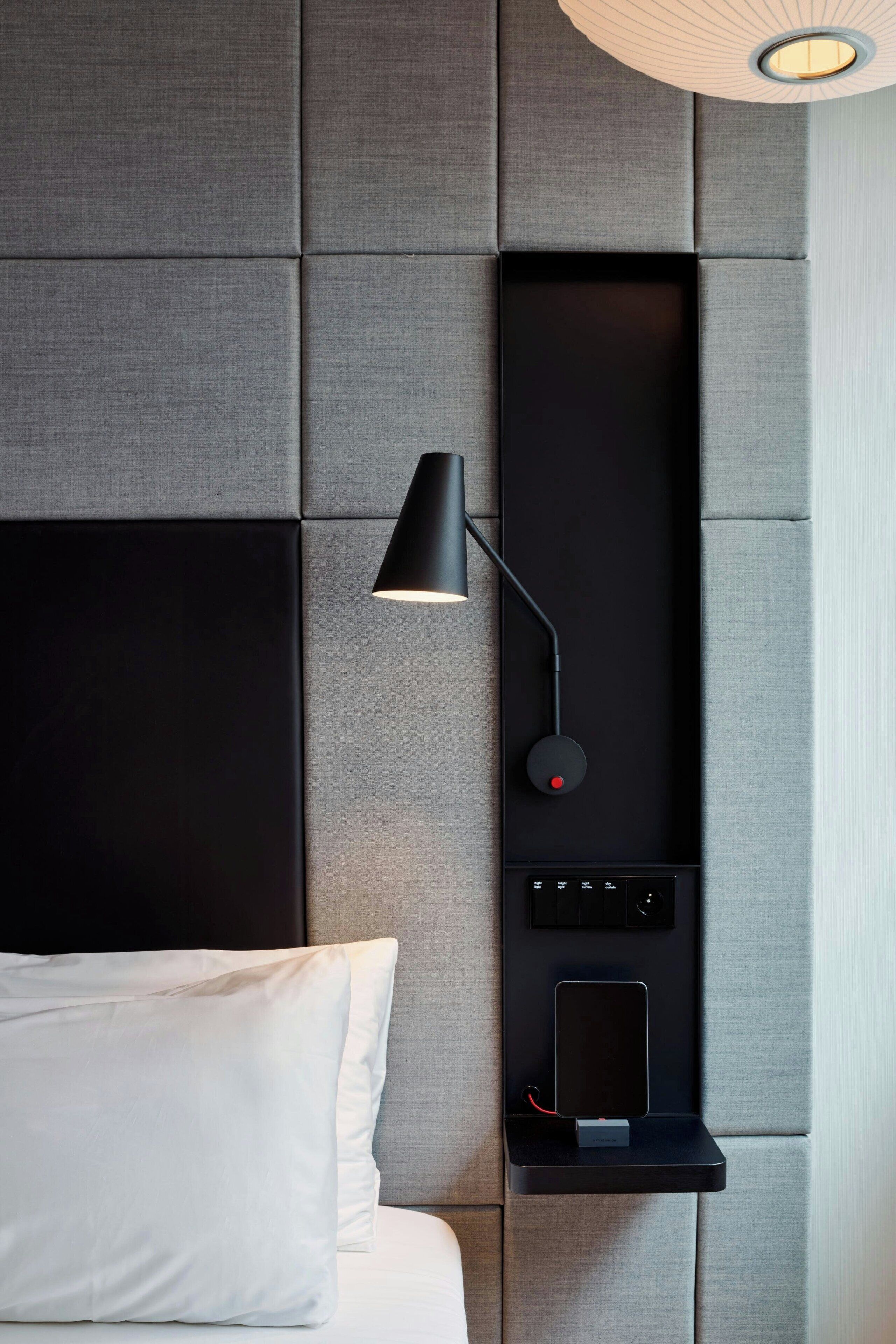 A modern bedroom with a large bed, a wall-mounted lamp, and a built-in shelf with a tablet.
