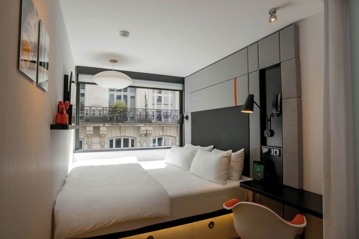 A modern hotel room with a large bed, a desk, and a view of a building through a window.