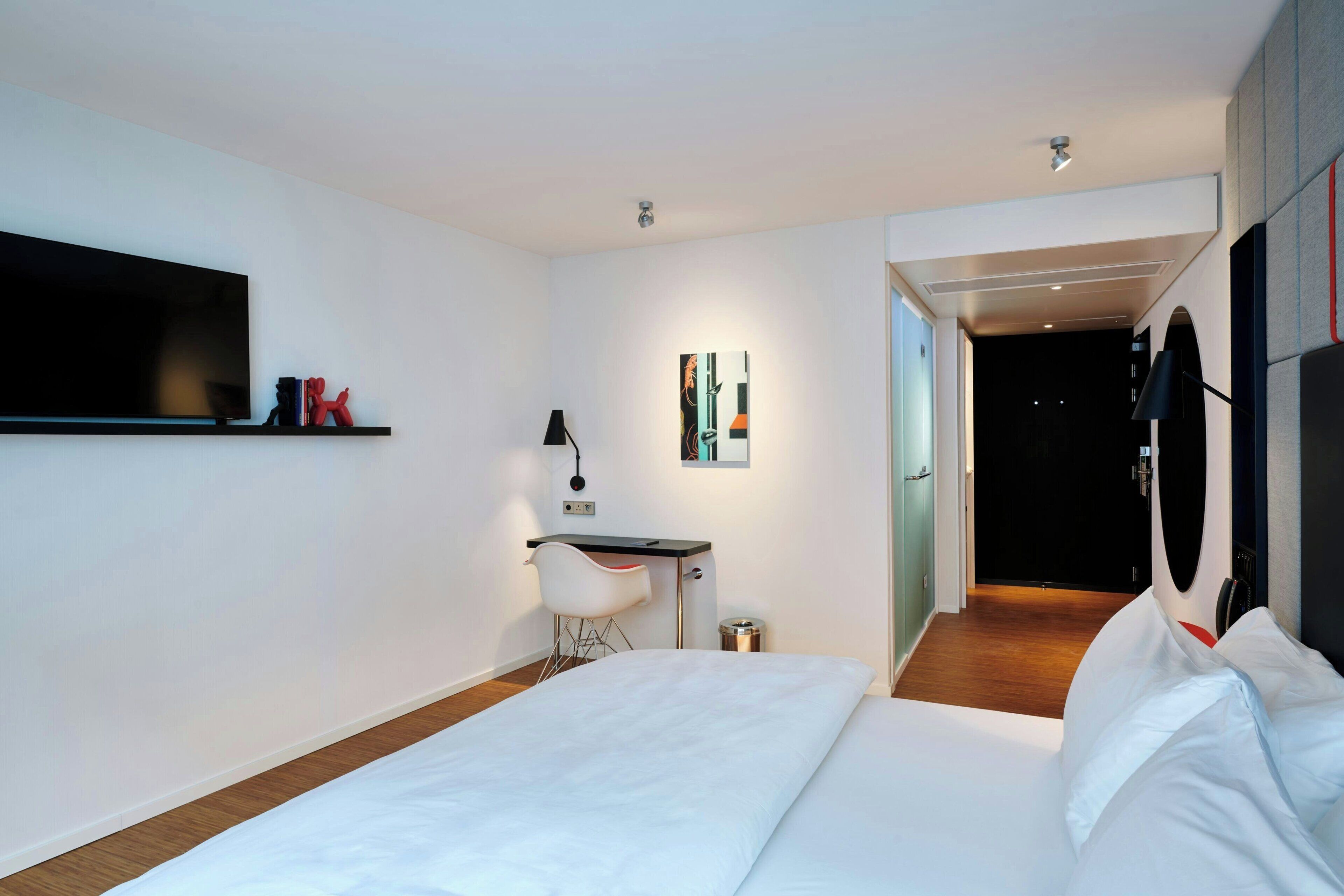 A modern hotel room with a large bed, a desk with a chair, a TV, and a painting on the wall.