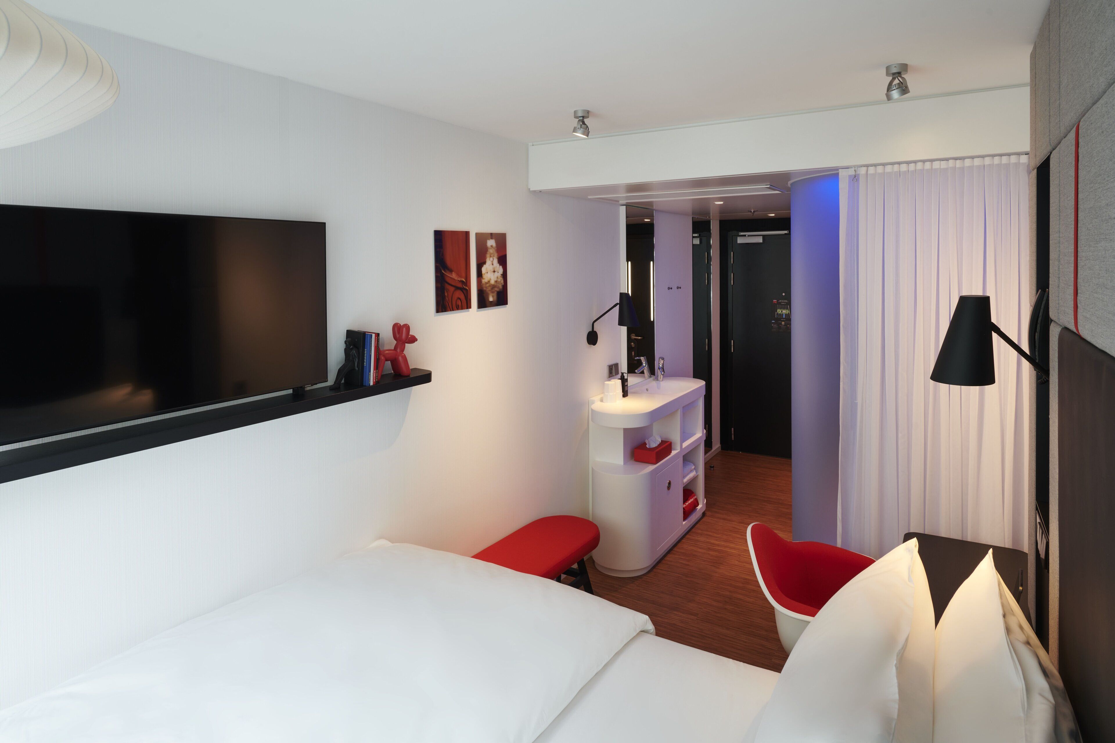 Een moderne hotelkamer met een grote flatscreen televisie, een wit bed, een rode fauteuil, een klein wit bureau met een lamp, en een badkamer zichtbaar door een geopende deur.