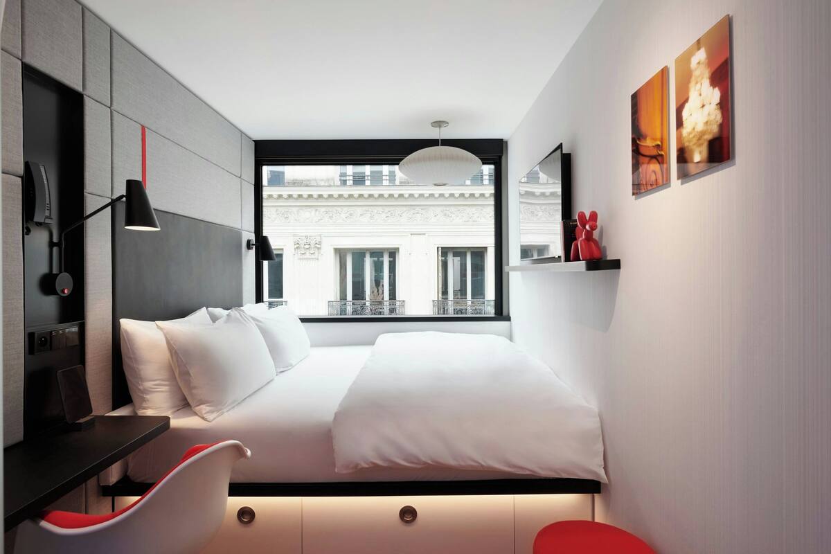 A modern hotel room with a bed, a desk, a chair, and a large window with a view of a building.