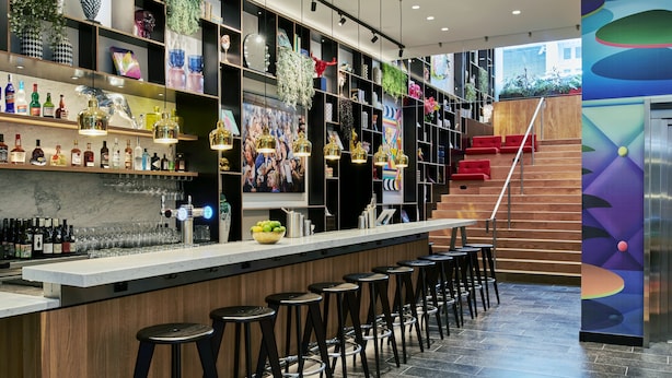 A modern bar area with a long counter, multiple stools, and shelves stocked with bottles and glasses.