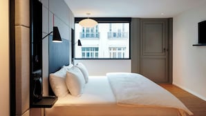 A modern bedroom with a bed, bedside lamps, and a view of buildings through a large window.
