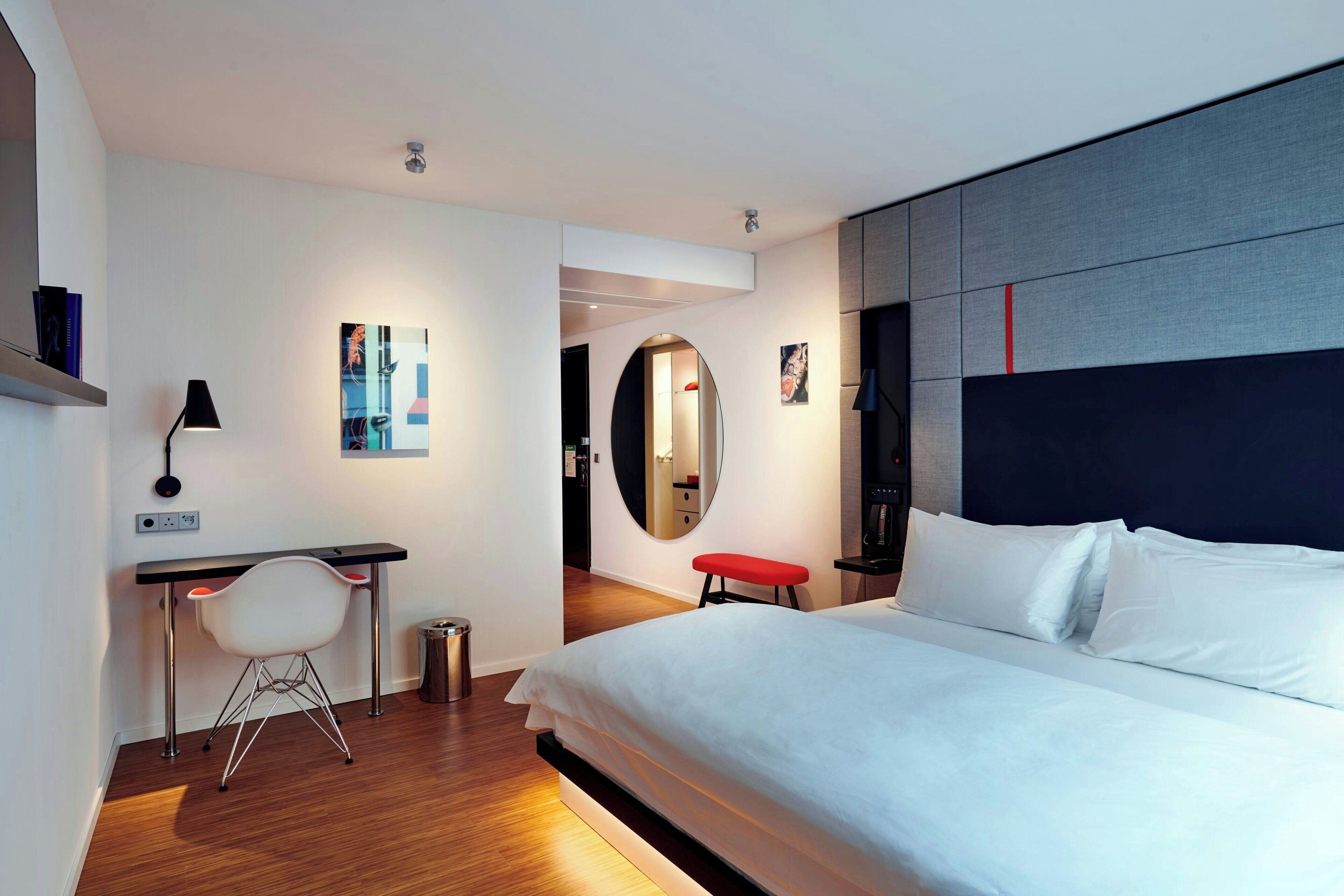 A modern hotel room with a large bed, a desk with a chair, a small stool, a mirror, and artwork on the walls.