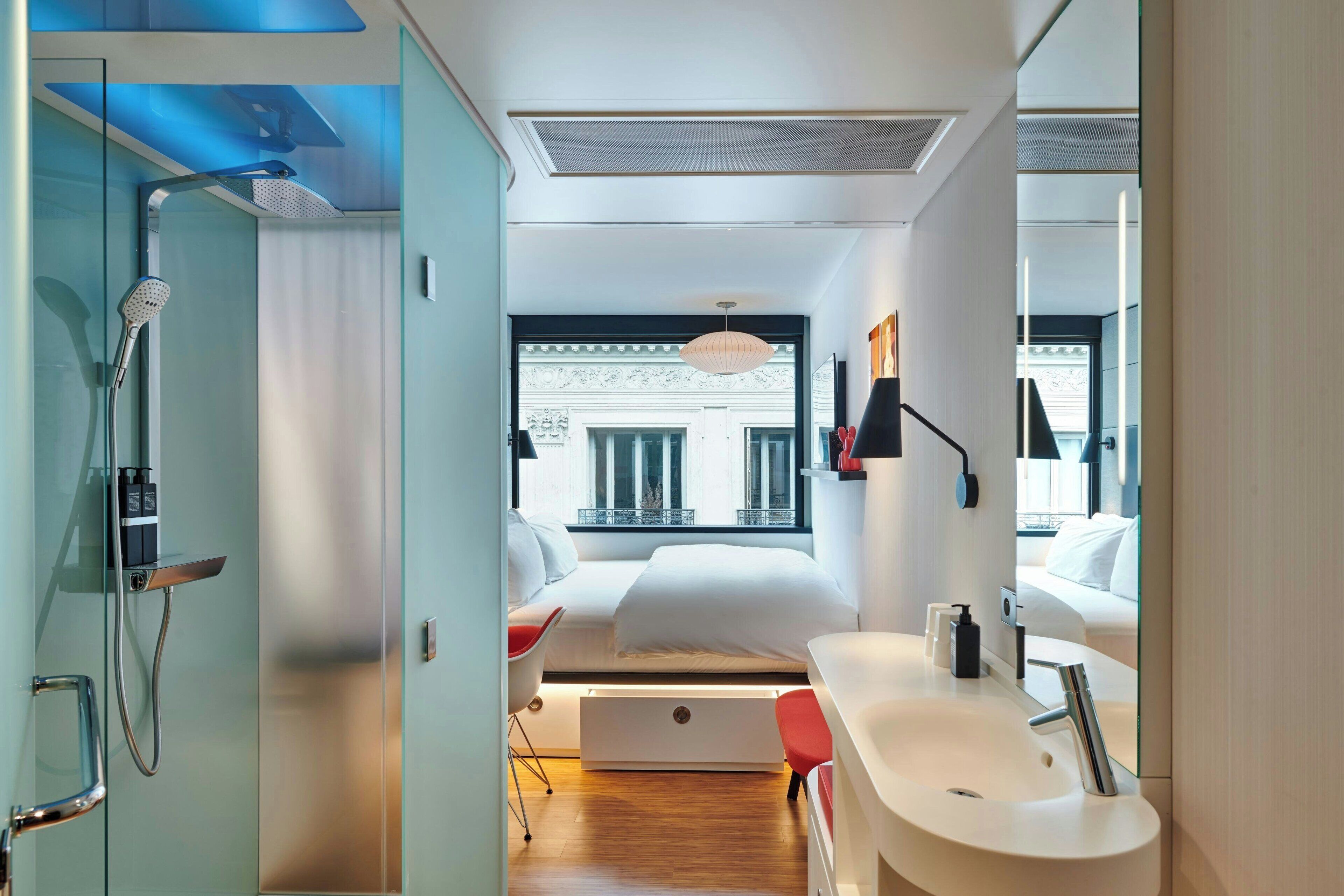 A modern hotel room with a shower, a bed, a bedside table, a sink, and a window with a view of a building.