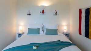 Een net opgemaakt bed met een blauwgroene deken en kussens, twee nachtlampjes en wanddecoratie met zeilboten.