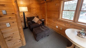 Acogedora habitación tipo cabaña con paredes de madera, cama con almohadas, una mesita con lámpara y ventana con vista a árboles.