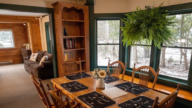 Un comedor acogedor con una mesa de madera dispuesta para cuatro personas, rodeada de sillas de madera. Se ve una estantería llena de libros al fondo.