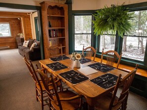 Un comedor acogedor con una mesa de madera dispuesta para cuatro personas, rodeada de sillas de madera. Se ve una estantería llena de libros al fondo.