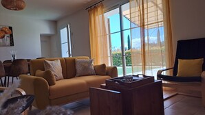 Un salon avec un canapé, une table basse et un fauteuil. Il y a une fenêtre avec des rideaux, une porte et un luminaire fixé au mur.
