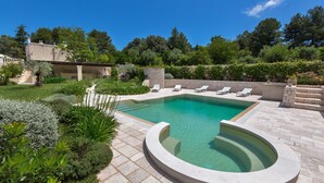 Une piscine à bord incurvé, entourée d’une terrasse en pierre et d’un jardin luxuriant où poussent différentes espèces de plantes, le tout délimité par un mur de pierre.
