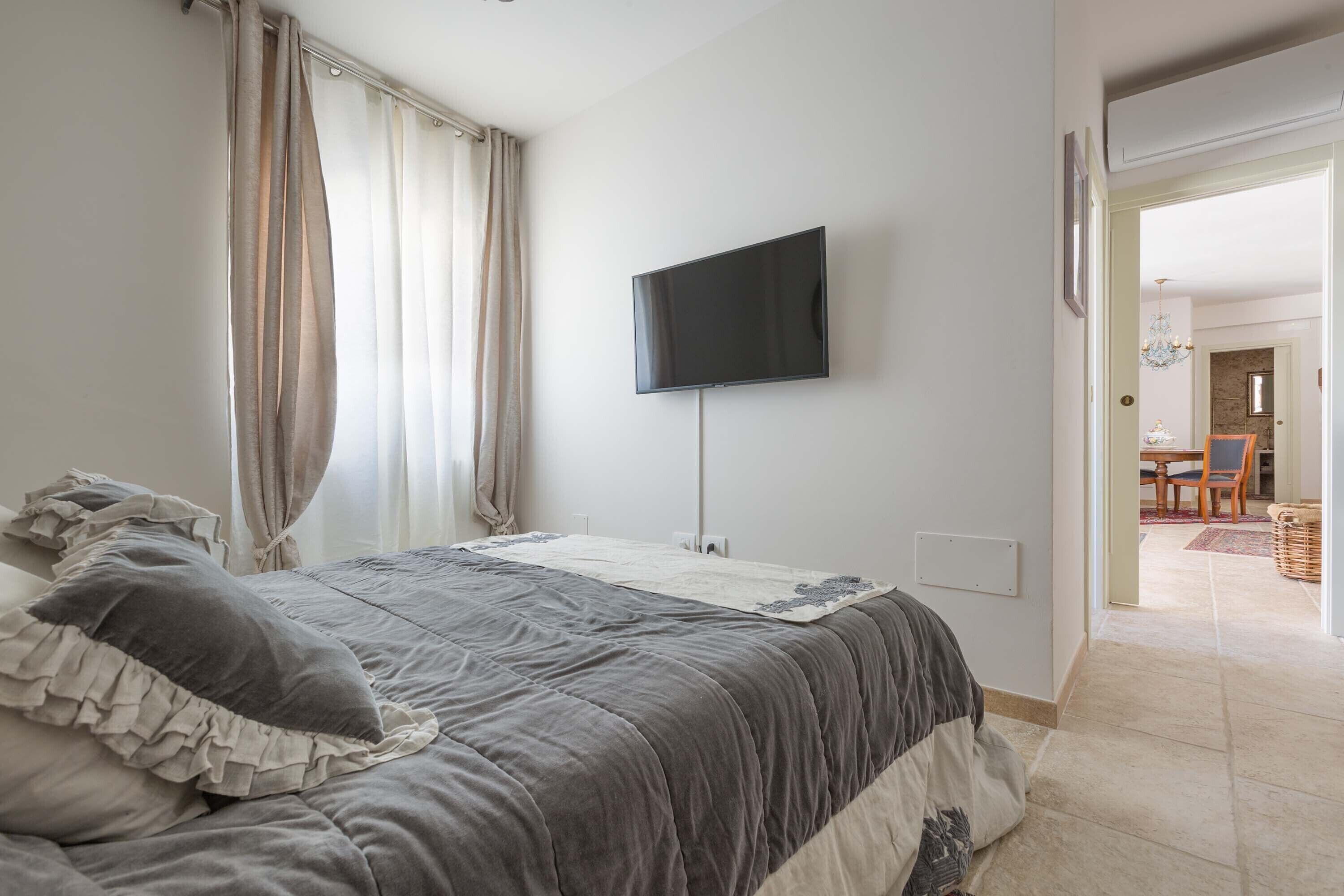 A bedroom with a bed, curtains, a flat-screen TV, and a view of a dining area.