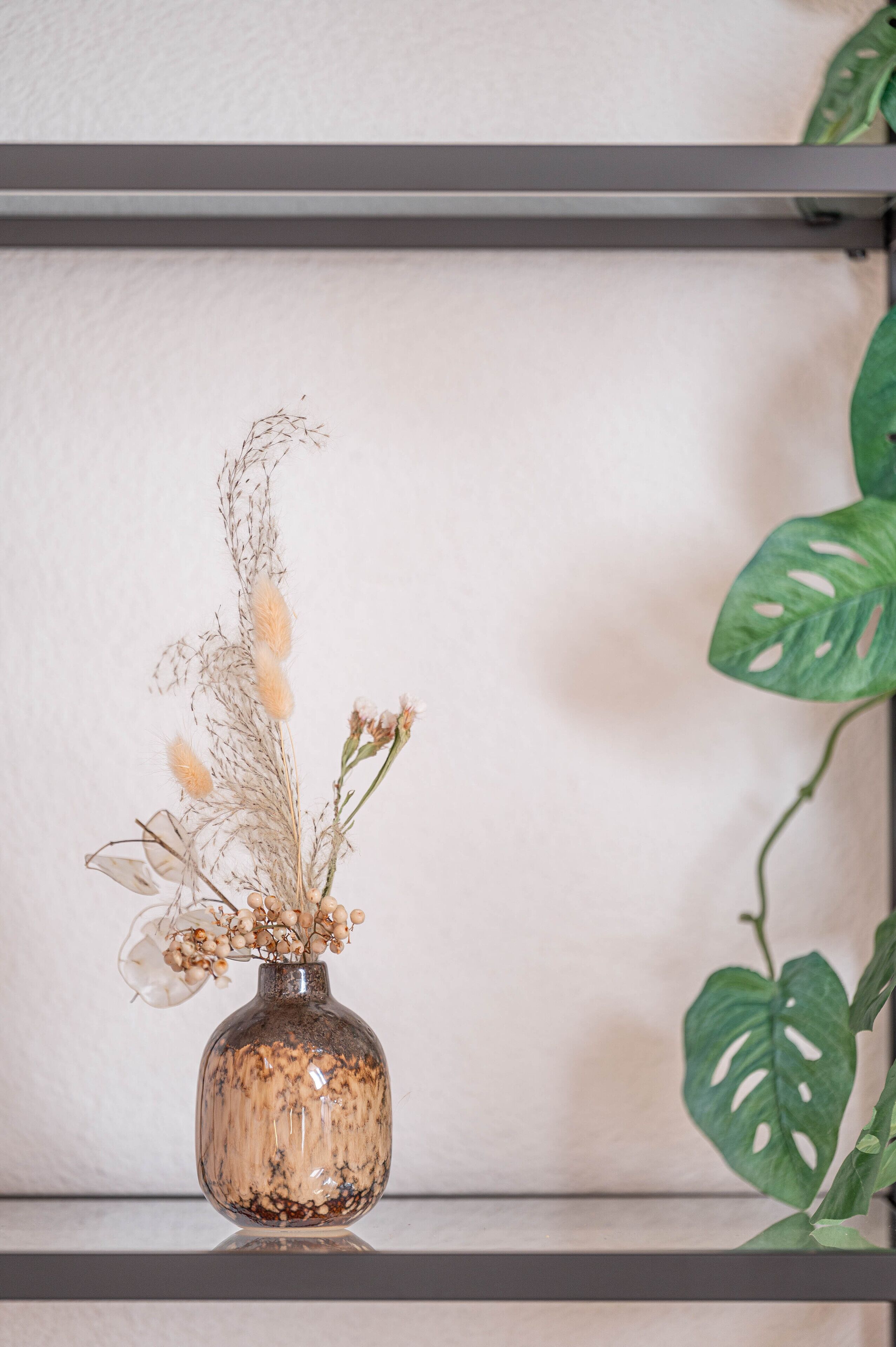 Eine transparente Glasvase mit getrockneten Blumen und Grünpflanzen auf einem Regal vor einer schlichten Wand.