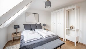 Ein Schlafzimmer mit schrägen Wänden, einem Bett mit grauer Bettwäsche, zwei Nachttischlampen, einem Nachttisch mit Lampe, einer kleinen Pflanze und einem weißen Kleiderschrank.