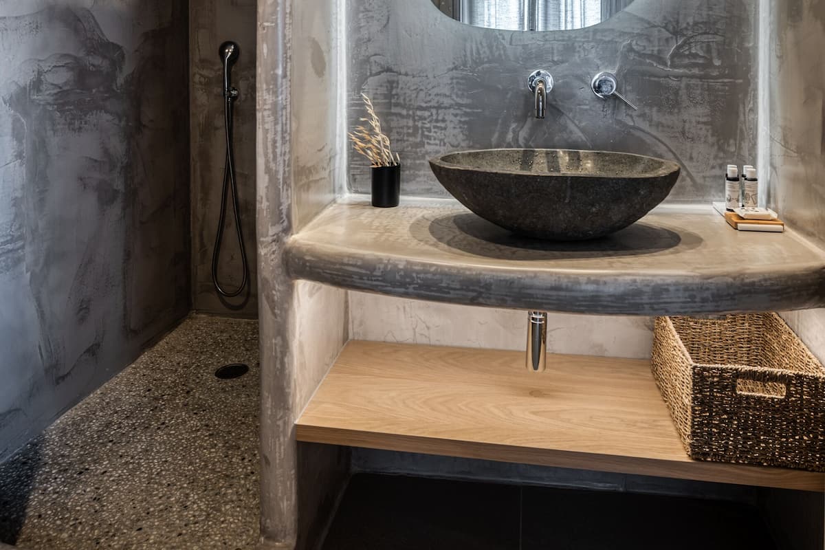 Uma casa de banho moderna com uma grande lavatório de pedra escura e arredondada, um móvel de banheira em madeira, chuveiro com porta de vidro e um espelho grande com moldura circular.