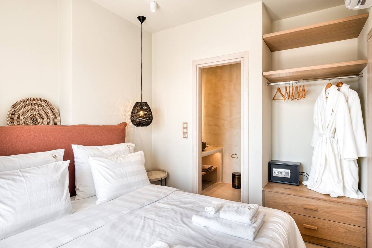 Quarto de hotel com cama, mesa de cabeceira, abajur, armário com roupas penduradas e prateleira com toalhas.