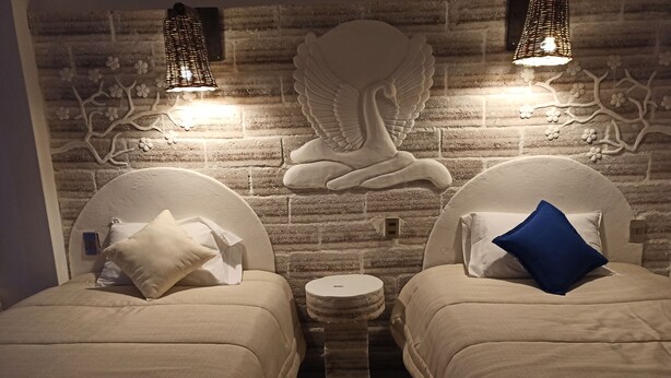 A room with two beds, each with a headboard and a pillow. There is a small round table between the beds. The wall behind the beds features a brick pattern and decorative elements.